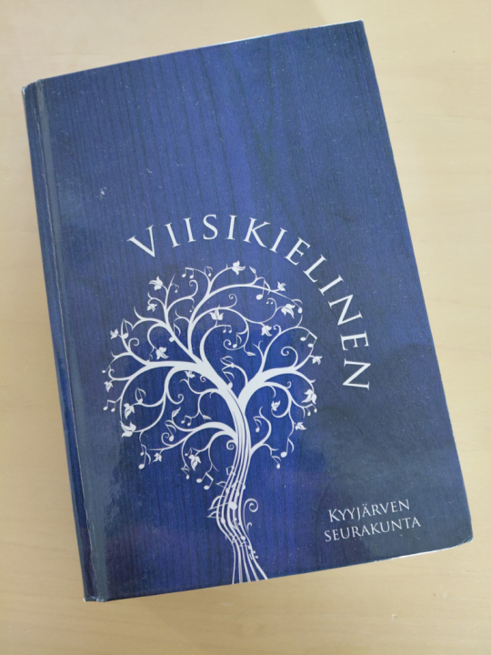 Kuvassa kirkon viidennen herätysliikkeen laulukirja, sinikantinen Viisikielinen, jossa kirjan nimi ja ja piirretty puu valkoisella. Puun runko alkaa nuottiviivastona., jossa myös muutama nuotti.