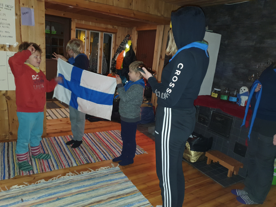 Etualalla sudenpentu antaa partiolupausta, jonka vastaanottaa nuori ryhmänjohtaja. Taustalla kaksi lasta, jotka pitelevät suomen lippua.