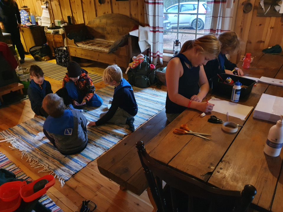 Lapset istuvat lattialla pelaamassa korttia, toiset lapset istuvat pöydän ääressä piirtämäss.